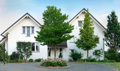 Two houses with roof gables are connected. In front of the house is a large round paved driveway. A flower island with roses. © Tanja Esser
