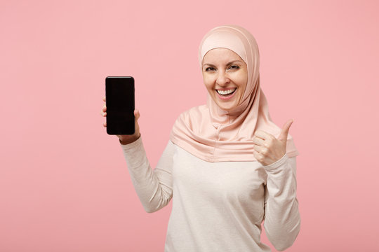 Arabian muslim woman in hijab light clothes posing isolated on pink  background. People religious Islam lifestyle concept. Mock up copy space.  Hold cellphone with blank empty screen, showing thumb up. Stock Photo