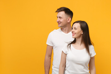 Beautiful smiling young couple two friends guy girl in white empty blank design t-shirts posing isolated on yellow orange wall background. People lifestyle concept. Mock up copy space. Looking aside.