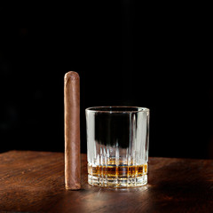 Strong alcoholic drink, scotch whisky or cognac in old fashion crystal glass  with smoking cigar  next to it on wooden table with black background