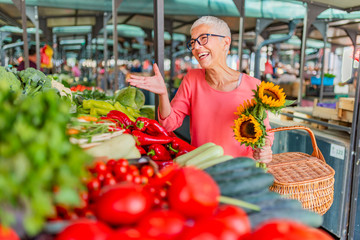 Woman buying vegetables at farmer's market stall. Basket full of vegetables and fruits on open market. Healthy mature woman shopping farmers market fresh fruits and vegetables