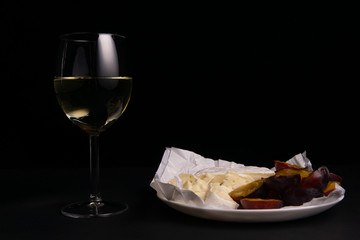 glass of wine next to a slice of cheese, plums on a black background