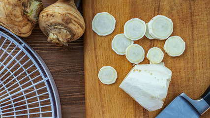 Chopped pieces of root parsnip lie on a cutting board next to a kitchen knife, a round form for drying and two fresh parsnip roots, top view