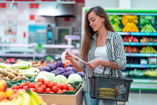 Young woman with shopping basket checks and examines a sales receipt after purchasing food in a grocery store. Customer buying products at supermarket