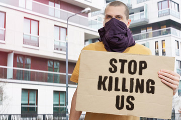 Man with covered face holds banner with an inscription: Stop Killing Us. Protestor holds a cardboard
