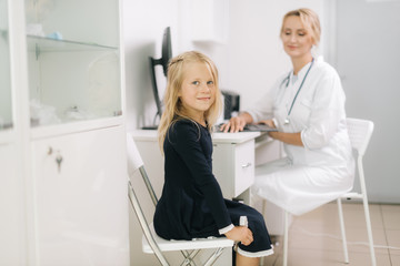 Cute kid girl with blond hair plays looking at the camera in pediatrician appointment. Doctor takes notes on a patient outpatient card.