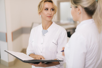 Fototapeta premium Two good-looking young female doctor looking at clipboard and working on patient record while standing in the corridor next