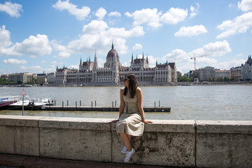 budapest travel: brunette girl in dress sits on the Danube embankment opposite the Hungarian...