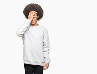 Young african american man with afro hair wearing sporty sweatshirt peeking in shock covering face and eyes with hand, looking through fingers with embarrassed expression.