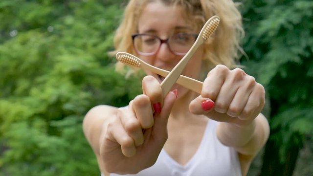 Young beautiful girl with ponytail is holding a useful bamboo toothbrushes outdoors during rainy weather on green tree background. Environmental friendliness and zero waste concept.