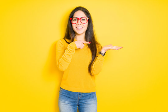 Beautiful brunette woman wearing red glasses over yellow isolated background amazed and smiling to the camera while presenting with hand and pointing with finger.