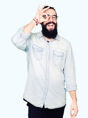 Young hipster man with long hair and beard wearing glasses doing ok gesture with hand smiling, eye looking through fingers with happy face.