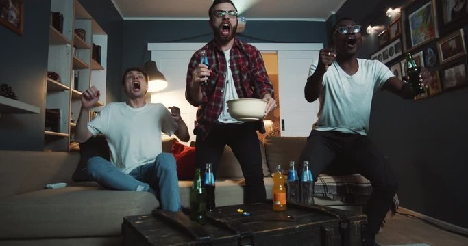 Happy diverse male sports fans get wild celebrating goal while watching game on TV at home with drinks slow motion.