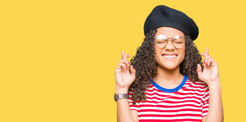 Young beautiful woman with curly hair wearing glasses and fashion beret smiling crossing fingers...
