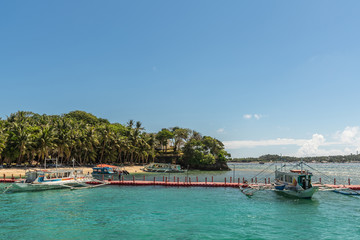 Fototapeta na wymiar Manoc-Manoc, Boracay, Philippines - March 4, 2019: Cagban Jetty Port and floating pier with small outrigger tourist ferries vessels on azure sea, under blue sky with green horizon foliage.