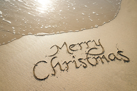 Merry Christmas message handwritten in smooth sand with an oncoming wave on the shore of a tropical beach