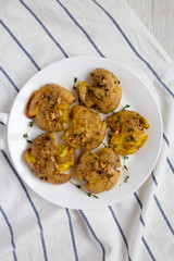 Homemade garlic thyme smashed potatoes on a white plate on a white wooden surface, top view. Flat lay, overhead, from above. Copy space.