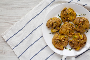 Homemade garlic thyme smashed potatoes on a white plate on a white wooden surface, top view. Flat lay, overhead, from above. Copy space.
