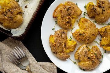 Homemade garlic thyme smashed potatoes on a white plate on a black background, top view. Flat lay, overhead, from above. Close-up.
