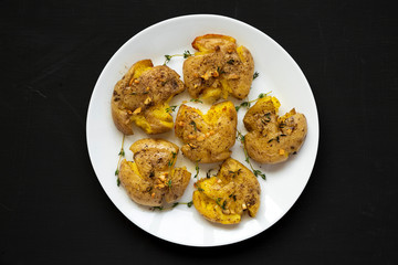 Homemade garlic thyme smashed potatoes on a white plate on a black surface, top view. Flat lay, overhead, from above. Close-up.