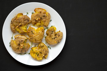 Homemade garlic thyme smashed potatoes on a white plate on a black background, top view. Flat lay, overhead, from above. Space for text.