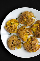 Homemade garlic thyme smashed potatoes on a white plate on a black background, top view. Flat lay, overhead, from above. Close-up.