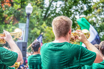 Young clean cut men in sweaty green t shirts play trumpets in parade with bokeh trees and American...