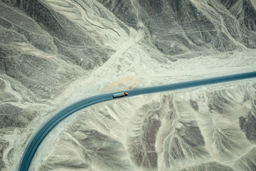 Aerial view looking down over a large orange and grey truck driving along a long rural road through the desert of Peru.