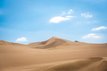 Fototapeta na wymiar The swirling sand dunes of the Peruvian desert with light fluffy clouds above on a warm sunny day creating a spectacular contrast across the horizon