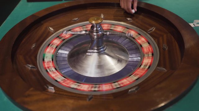 A man spinning a Roulette wheel and dropping the ball into the wheel. Waiting for it to land on a lucky number.