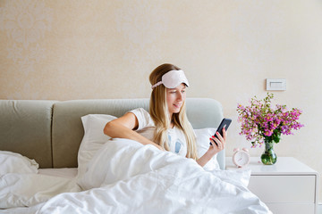 Happy young blonde woman in pajamas and blindfold waking up and using mobile phone in bed, reading news, working or watching social media. Morning light in bedroom. People using technology concept