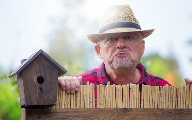 An elderly man with hat looks angry and watching over a garden fence. Concept problems with the...