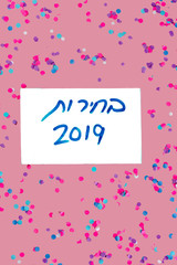 Hebrew text Elections 2019 on white isolated voting paper over pink with colorful confetti background. Israeli legislative Elections for Knesset Israel 2019.