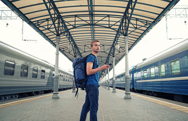 Tourist backpacker with smartphone stand on railway station platform and waiting for train. Travel concept.