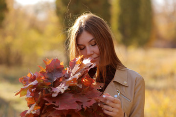 Woman in the autumn sun with yellow maple leaves. Girl holding a bouquet of autumn leaves at sunset in a forest.