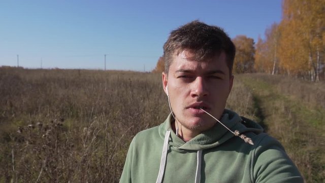 Handsome caucasian young man with thin stalk in mouth, wearing light-green hoody, is slowly walking alone in countryside along empty field on clear autumn day with blue sky.