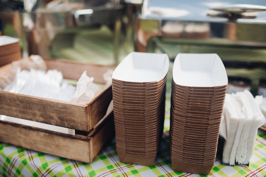 Stock photo of paper recyclable containers in stacks on the table over patterned green and white tablecloth. Box of napkins and utensils. Outdoor food festival.