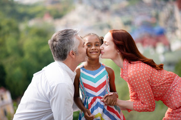 Fototapeta premium Adoptive parents kissing their cute dark-skinned girl
