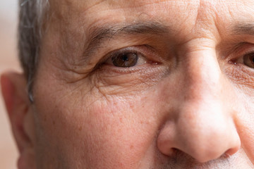 A close up view on the eye and nose of an elderly man, with gray hair and eyebrows, aging process of the skin is seen in detail with wrinkles and crow's feet.