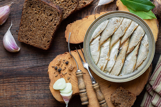 Smoked sprat in a tin served with rye bread and garlic cloves on a rustic wooden cutting board. Overhead view, flat lay