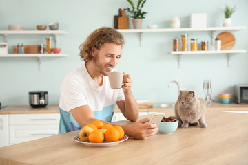 Man with mobile phone drinking coffee while sitting in kitchen with cute cat - Powered by Adobe