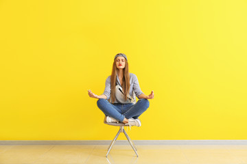 Beautiful young woman meditating on chair near color wall