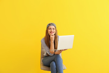 Happy young woman with laptop sitting on chair against color background