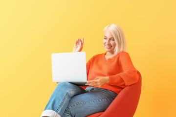 Mature woman with laptop sitting on chair against color background