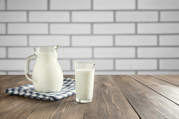 Glass of fresh milk and jug on wooden tabletop and white brick wall as background with space for display product.