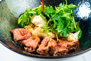Warm salad with roast beef, crackers, arugula and sauce in a black bowl on a marble table. Tasty and healthy snack for lunch. Food photo background for menu or recipe. Restaurant