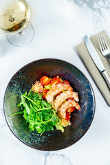 Salad with shrimp, guacamole, arugula and cherry tomatoes in a stylish dark bowl on a marble table with white wine. Seafood salad. Food photo for menu or recipe. Top view, flat lay