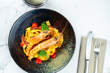 Homemade Italian pasta with cherry tomatoes and seafood. Pasta with fresh and tasty langoustines shrimp in a dark stylish bowl on a marble table. Food photo for menu or recipe. Restaurant
