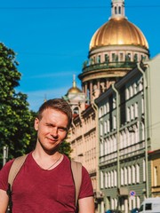 A blond man in a pink t-shirt and with a backpack stands against the background of St. Isaac's Cathedral in St. Petersburg