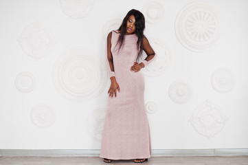 Fashionable african american model woman in pink brilliant evening dress posed against white decorative wall.